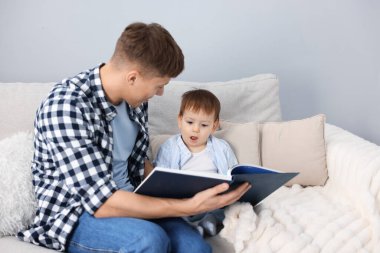 Erkek dadı, evdeki gri duvarın yanındaki kanepede oturan şirin çocuğa kitap okuyor.