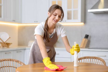 Temizlik malzemesi ve mikrofiber bez taşıyan bir kadın mutfaktaki ahşap masayı siliyor.