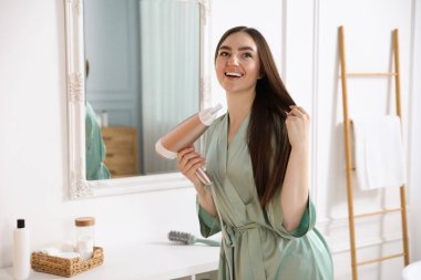 Bornozlu gülümseyen kadın saçını banyoda saç kurutma makinesiyle kurutuyor.