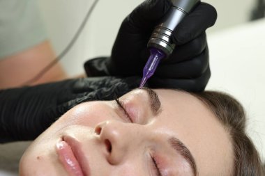 Güzellik uzmanı, içeride genç bir kadına kalıcı kaş makyajı yapıyor.