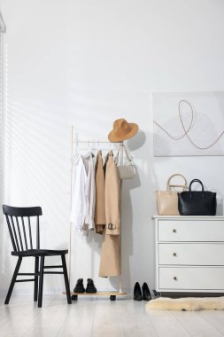 Askıda asılı farklı kıyafetler, aksesuarlar, ayakkabılar ve beyaz duvarın yanındaki sandalye.