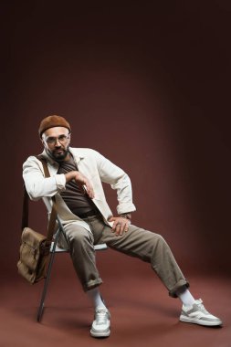 Moda modeli. Kahverengi arka planda, sandalyede oturan yakışıklı adam.
