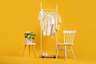 Askıda asılı farklı kıyafetler, kep, ayakkabı, çiçek ve sandalye turuncu arka planda