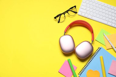 Kulaklıklar, bilgisayar klavyesi, sarı arka planda gözlük ve kırtasiye malzemesi, düz konum. Metin için boşluk