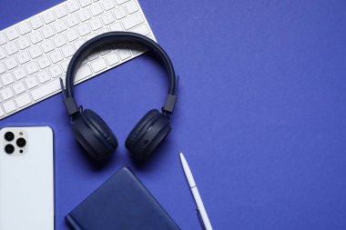 Kulaklıklar, bilgisayar klavyesi, akıllı telefon ve mavi arka planda kırtasiye malzemesi, düz konum. Metin için boşluk