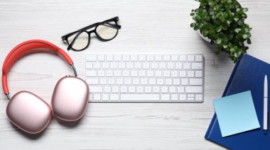 Kulaklıklar, bilgisayar klavyesi, gözlükler, çiçek dekoru ve beyaz ahşap masa üzerindeki kırtasiye malzemesi, düz konum