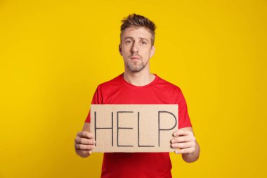 Mutsuz adamın elinde kelime yazılı karton tabela var. Sarı arka planda yardım.