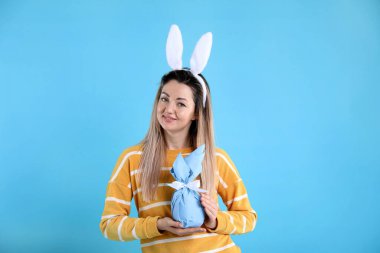 Paskalya kutlaması. Saç bandı takmış, tavşan kulaklı, gülümseyen bir kadın açık mavi arka planda hediyesini tutuyor.