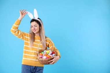 Paskalya kutlaması. Saç bandı takmış, tavşan kulaklı ve açık mavi arka planda boyalı yumurta dolu sepetli gülümseyen bir kadın.