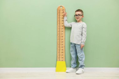 Küçük çocuk, açık yeşil duvarın yanında çizelgesiyle boyunu ölçüyor.