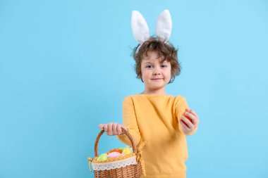 Easter celebration. Cute little boy in headband with bunny ears and wicker basket full of painted eggs on light blue background. Space for text