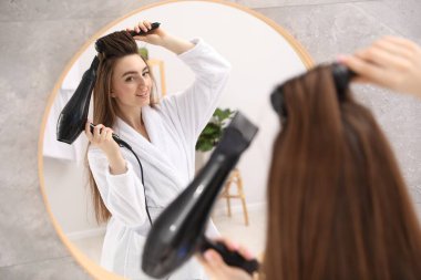 Bornozlu gülümseyen kadın saçını saç kurutma makinesiyle şekillendiriyor ve banyoda aynanın yanında tarıyor.