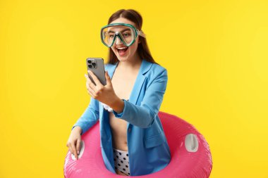 Sarı arka planda şişme yüzük, yüzme maskesi ve akıllı telefonu olan bir iş kadını.