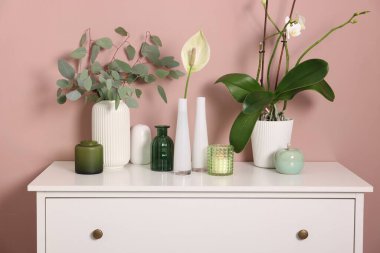 Koyu bej duvarın yanındaki dolapta çiçek dekoru ve yanan mum olan farklı güzel vazolar.