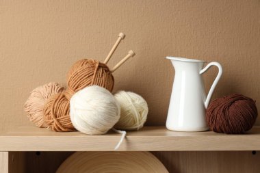 Bej duvarın yanındaki rafta örgü iğneleri, iplikler ve sürahiler var.