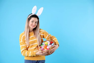 Paskalya kutlaması. Saç bandı takmış, tavşan kulaklı ve açık mavi arka planda boyalı yumurta dolu sepetli gülümseyen bir kadın.