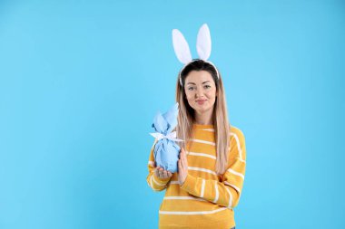 Paskalya kutlaması. Saç bandı takmış, tavşan kulaklı güzel bir kadın açık mavi arka planda hediye tutuyor, mesaj için yer var.