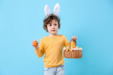 Paskalya kutlaması. Saç bantlı, tavşan kulaklı ve açık mavi arka planda boyalı yumurtalarla dolu sepeti olan sevimli bir çocuk.