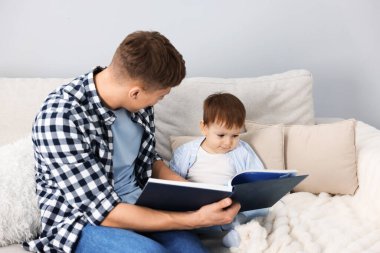 Erkek dadı, evdeki gri duvarın yanındaki kanepede oturan şirin çocuğa kitap okuyor.