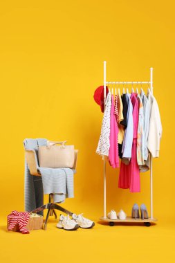 Askıda asılı farklı kıyafetler, aksesuarlar, ayakkabılar ve sandalye turuncu arka planda