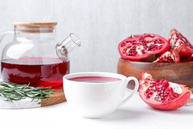 Aromatik nar çayı fincanda, çaydanlıkta, taze meyve parçaları ve biberiye beyaz masada, arka planda ışığa karşı, yakın plan