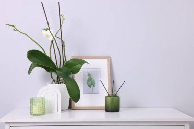 Güzel orkide saksıda çiçek, kamış oda spreyi, beyaz çekmecelerde resim ve mum ışık duvarı yakınında, yazı için yer