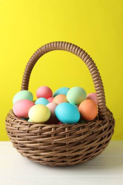 Güzel dekore edilmiş Paskalya yumurtaları hasır sepetin içinde beyaz ahşap masada sarı arka planda, yakın planda.