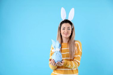 Paskalya kutlaması. Saçbandı takmış, tavşan kulaklı, açık mavi arka planda hediyesini tutan gülümseyen bir kadın. Mesaj için yer var.
