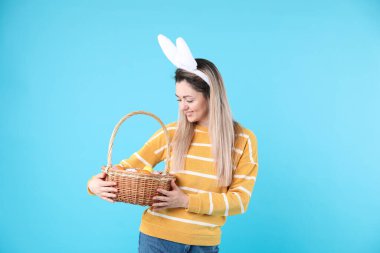 Paskalya kutlaması. Saçbantlı, tavşan kulaklı ve açık mavi arka planda boyalı yumurtalarla dolu sepeti olan gülümseyen kadın.