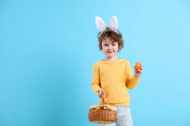 Paskalya kutlaması. Saçbantlı, tavşan kulaklı, açık mavi arka planda yumurta ve hasır sepetli gülümseyen bir çocuk. Metin için boşluk