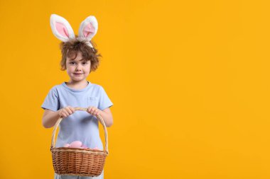 Easter celebration. Cute little boy in headband with bunny ears and wicker basket full of painted eggs on yellow background. Space for text