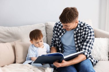 Gülümseyen erkek dadı, evdeki gri duvarın yanındaki kanepede oturan şirin çocuğa kitap okuyor.