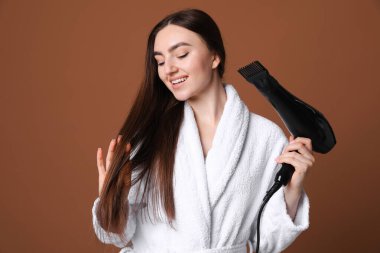 Bornozlu gülümseyen kadın saçını kurutuyor kahverengi arka planda saç kurutma makinesiyle.