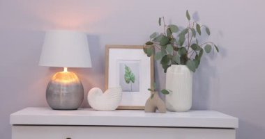 Okaliptüs dalları vazolarda, resim ve yanan mumlar gri duvarın yakınındaki beyaz çekmecelerde, kamera ileri doğru hareket ediyor.