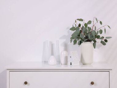 Okaliptüs dallı şık vazo ve ışık duvarı yakınındaki beyaz çekmecelerin üzerine dekor.