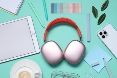Kulaklıklar, akıllı telefon, tablet, bardaklar, kahve, kırtasiye ve turkuaz arka planda yeşil yapraklar, düz konum