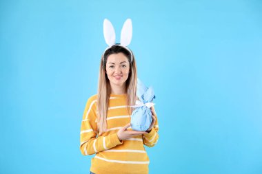 Paskalya kutlaması. Saç bandı takmış, tavşan kulaklı, gülümseyen bir kadın açık mavi arka planda hediyesini tutuyor.