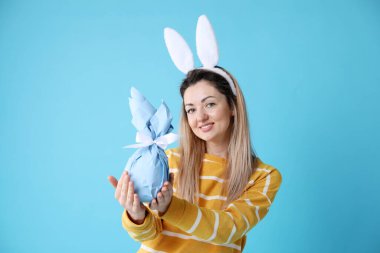 Paskalya kutlaması. Saç bandı takmış, tavşan kulaklı, gülümseyen bir kadın açık mavi arka planda hediyesini tutuyor.
