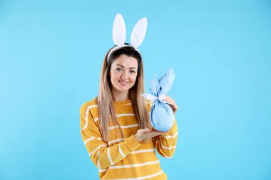 Paskalya kutlaması. Saç bandı takmış, tavşan kulaklı, gülümseyen bir kadın açık mavi arka planda hediyesini tutuyor.