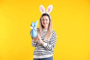 Paskalya kutlaması. Saç bandı takmış, tavşan kulaklı, gülümseyen bir kadın sarı arka planda hediyesini tutuyor.