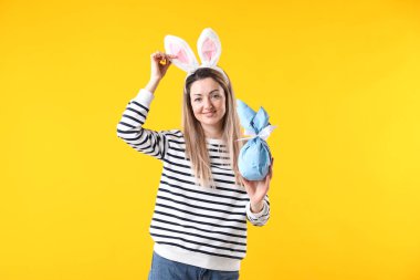 Paskalya kutlaması. Saç bandı takmış, tavşan kulaklı, gülümseyen bir kadın sarı arka planda hediyesini tutuyor.