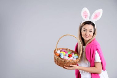 Paskalya kutlaması. Saç bandı takmış, tavşan kulaklı, gri arka planda yumurta dolu sepetli gülümseyen bir kadın. Mesaj için yer var.