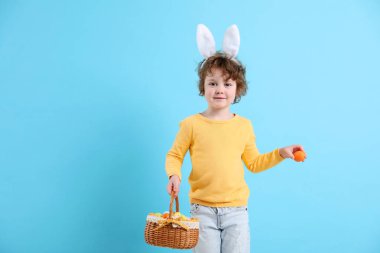 Paskalya kutlaması. Saç bantlı, tavşan kulaklı ve açık mavi arka planda boyalı yumurtalarla dolu sepeti olan şirin bir çocuk. Metin için boşluk