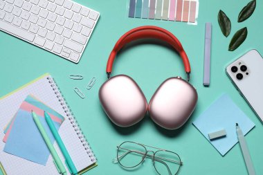 Kulaklık, akıllı telefon, gözlük, kırtasiye malzemesi ve turkuaz arka planda yeşil yapraklar.
