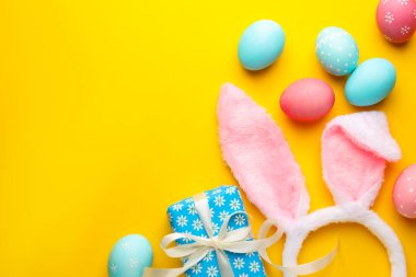 Boyalı Paskalya yumurtaları, tavşan kulaklığı ve sarı arka planda hediye, düz yatak. Metin için boşluk