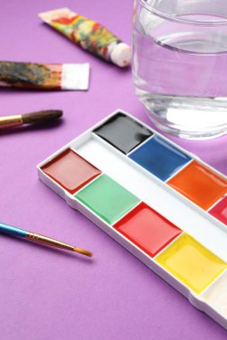 Mor zemin üzerinde suluboya boya, fırça, tüpler ve bir bardak su bulunan palet, yakın plan