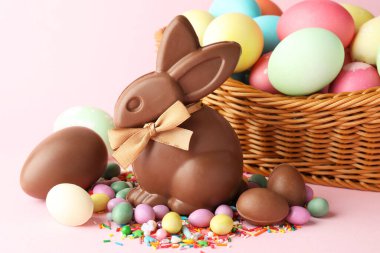 Çikolatalı Paskalya tavşanı, hasır sepette boyanmış yumurtalar, pembe arka planda şekerler ve serpiştirmeler, yakın plan.