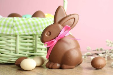 Çikolatalı Paskalya tavşanı ve hasır sepetteki yumurtalar ahşap masada pembe arka planda, yakın plan.