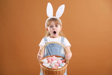 Paskalya kutlaması. Saçbantlı, tavşan kulaklı, kahverengi arka planda boyalı yumurta dolu sepeti olan duygusal küçük bir kızın portresi.