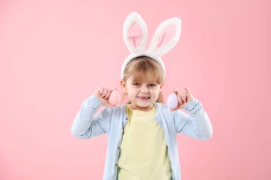 Paskalya kutlaması. Saçbantlı, tavşan kulaklı, pembe arka planda boyalı yumurta tutan küçük bir kızın portresi.